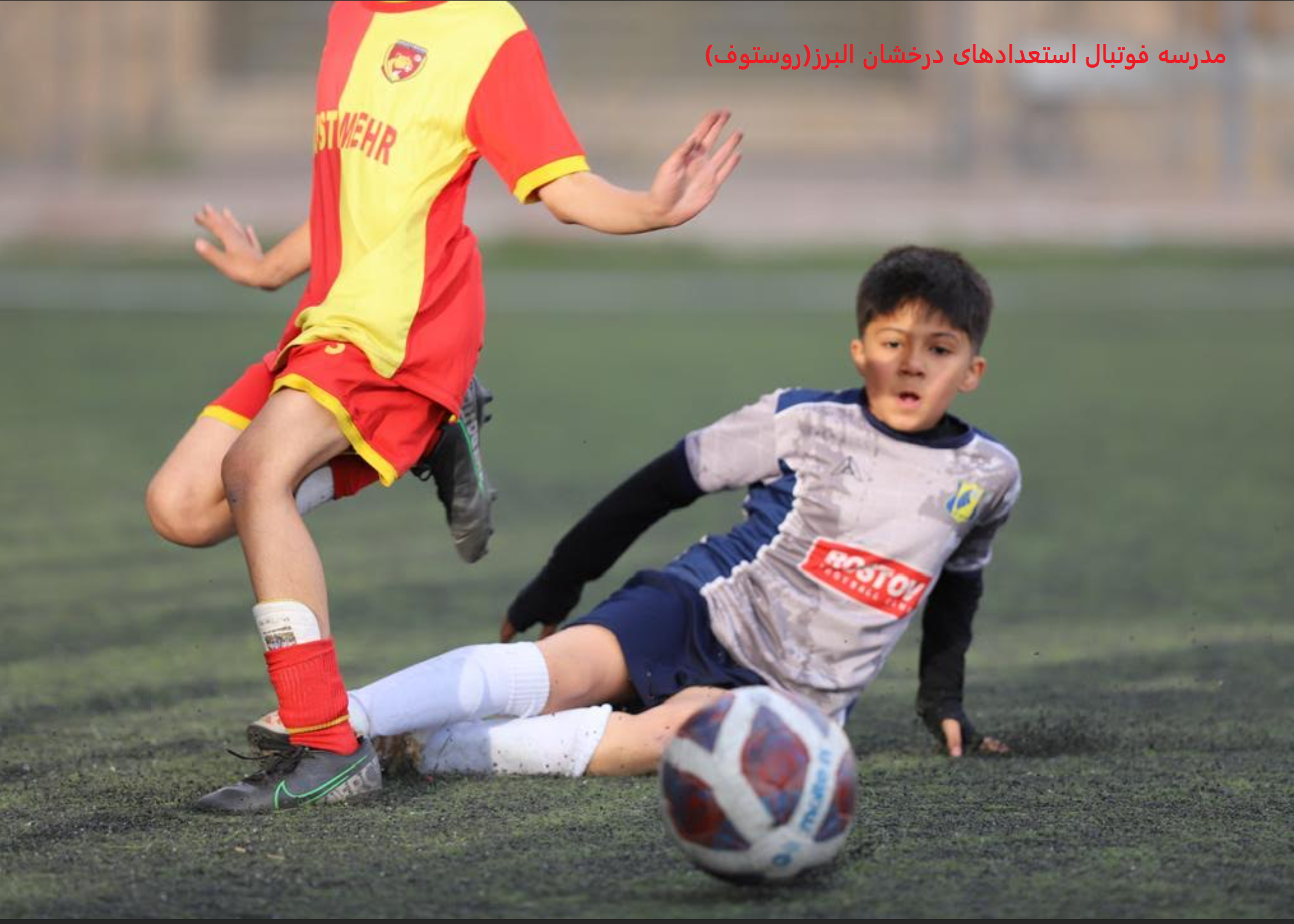 دو کودک در حال بازی فوتبال روی زمین چمن مصنوعی، یکی با لباس قرمز و زرد در حال دریبل کردن توپ و دیگری با لباس خاکستری و آبی که روی زمین افتاده است دو کودک در حال بازی فوتبال روی زمین چمن مصنوعی، یکی با لباس قرمز و زرد در حال دریبل کردن توپ و دیگری با لباس خاکستری و آبی که روی زمین افتاده است