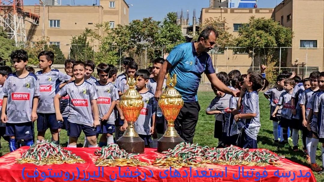 گروهی از کودکان با لباسهای ورزشی خاکستری و آبی با آرم 'روستوف' در حال ایستادن دور یک میز قرمز که دو جام طلایی و تعداد زیادی مدال و نشان روی آن قرار دارد، همراه با یک مربی که جامها را به آنها اهدا میکند، در محیطی سبز با ساختمانها در پسزمینه گروهی از کودکان با لباسهای ورزشی خاکستری و آبی با آرم 'روستوف' در حال ایستادن دور یک میز قرمز که دو جام طلایی و تعداد زیادی مدال و نشان روی آن قرار دارد، همراه با یک مربی که جامها را به آنها اهدا میکند، در محیطی سبز با ساختمانها در پسزمینه
